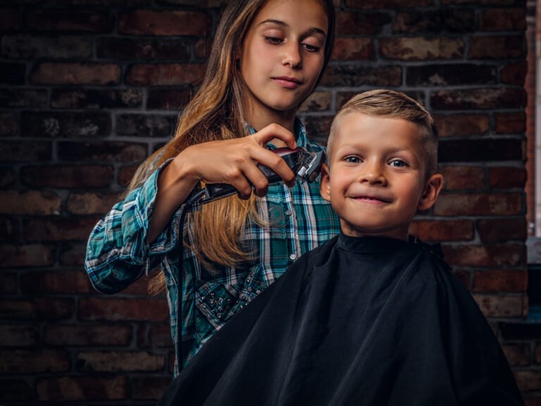 Smiling preschooler boy getting haircut. The older sister cuts her little brother with a trimmer against a brick wall.
